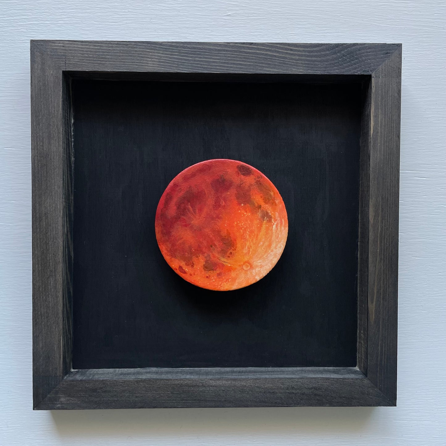 Black stained frame with black background. Floating in the shadowbox is a hand-painted clay Moon in orange