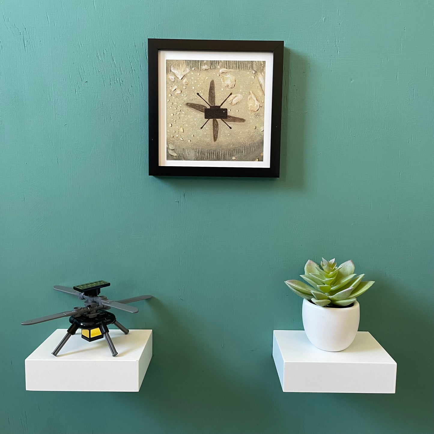 Zoomed out view of framed print. Below the frame are two small white shelves. One has a LEGO Ingenuity, one has a fake succulent.