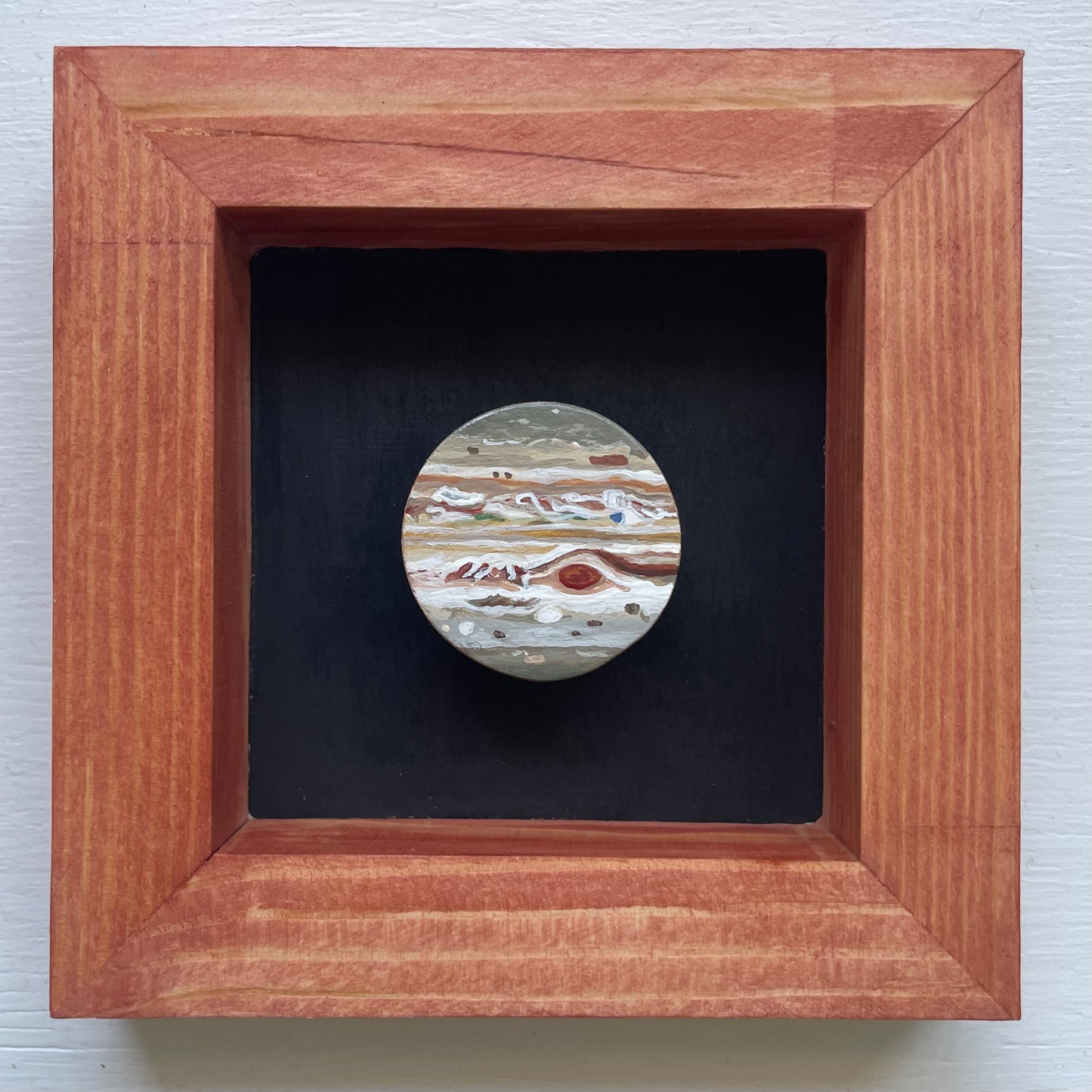 Red oxide stained frame with black background. Floating in the shadowbox is a hand-painted clay Jupiter