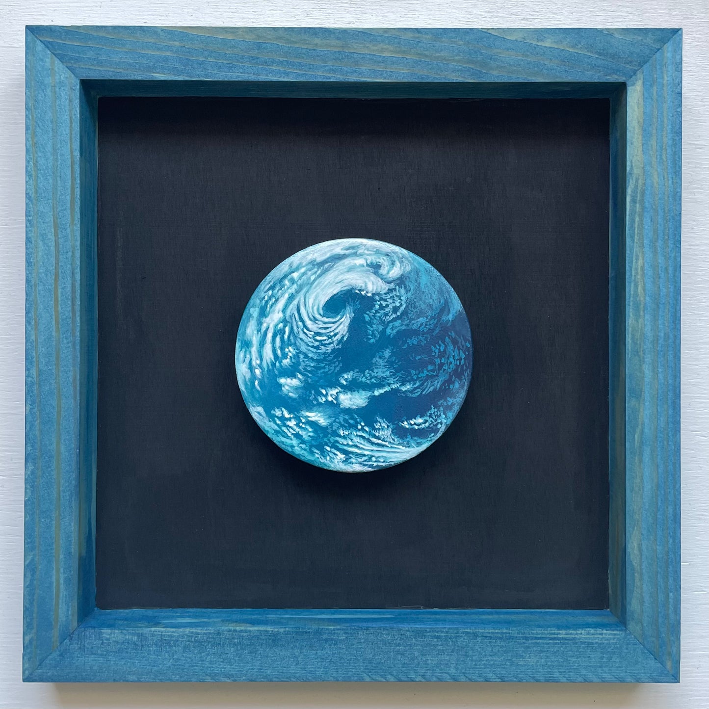 Medium blue stained frame with black background. Floating inside is a clay disk painted like a blue water world exoplanet with swirling white clouds