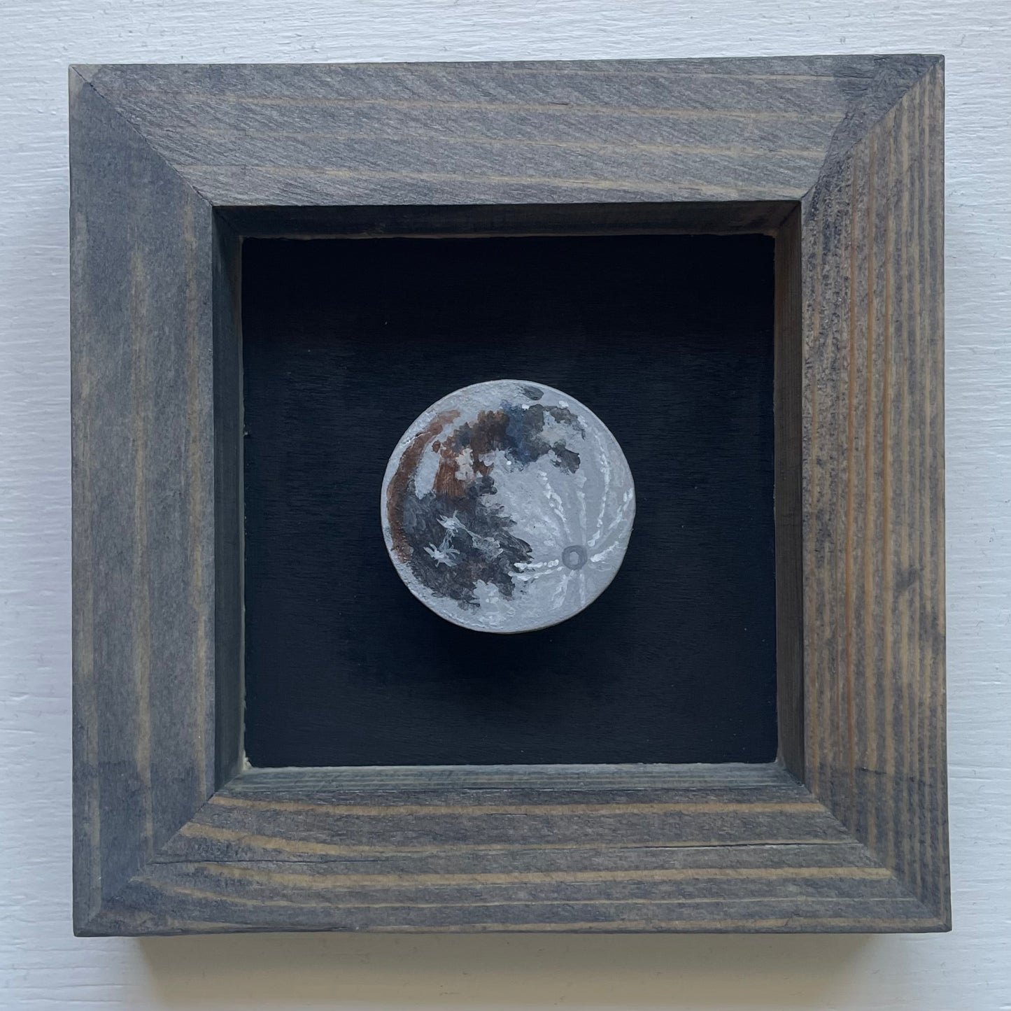 Grey stained frame with black background. Floating in the shadowbox is a hand-painted clay Moon