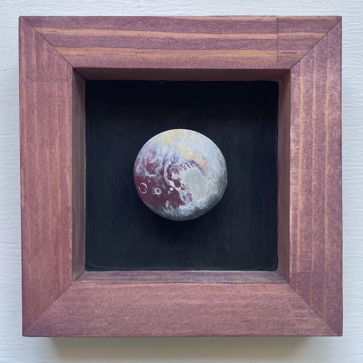 Deep reddish stained frame with black background. Floating in the shadowbox is a hand-painted clay Pluto