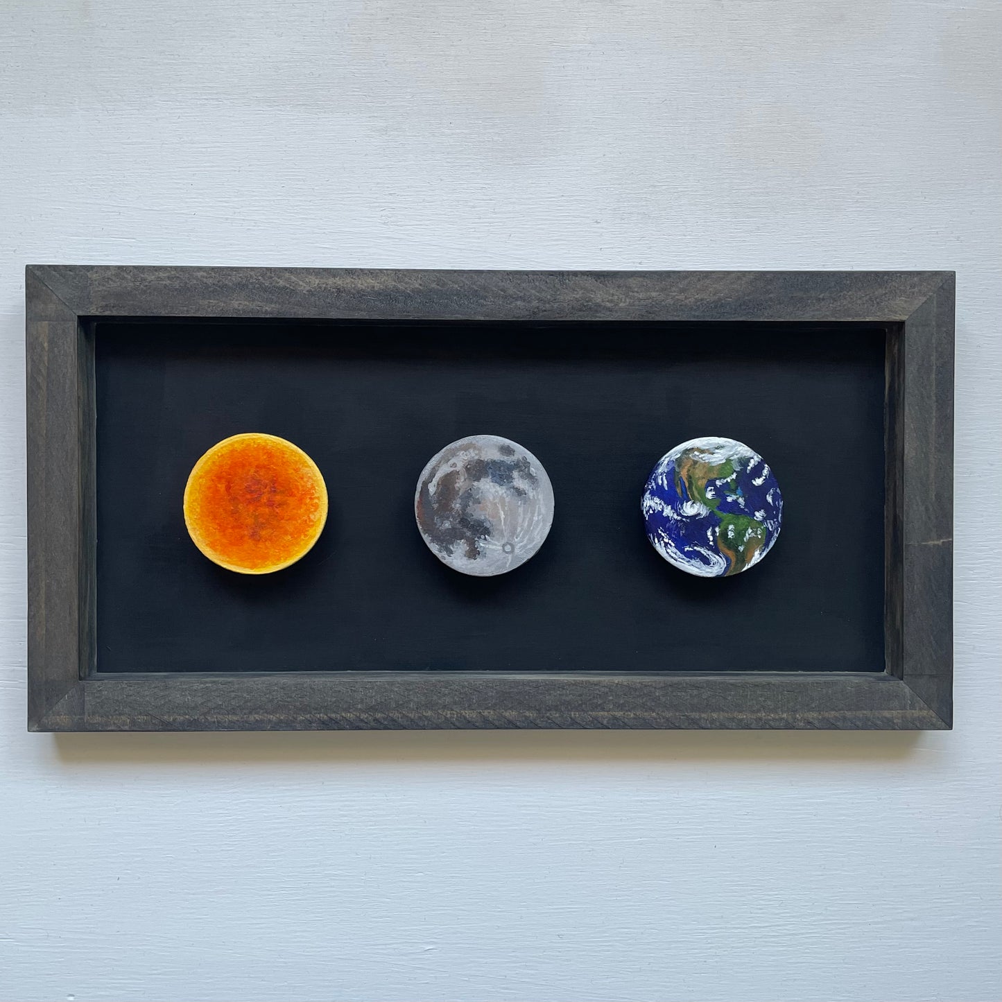Black stained frame with black background. Floating inside are three clay disks painted like the Sun, Moon, and Earth.