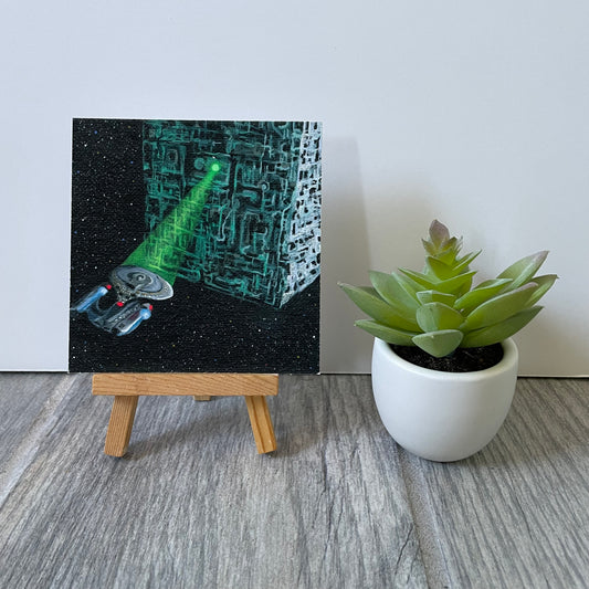 Mini print of starship and Borg cube on easel next to fake plant