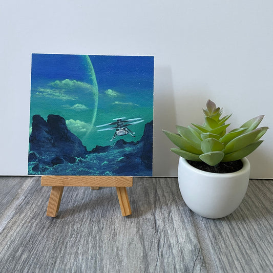 Mini print of a painting of a blue-green exoplanet surface with a little Ingenuity helicopter hovering. Painting is sitting on easel next to fake plant.