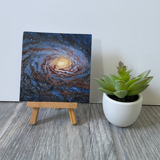 Mini painting of a blue and red-brown spiral galaxy. The center is yellow and orange. The painting is sitting on an easel next to a fake plant.