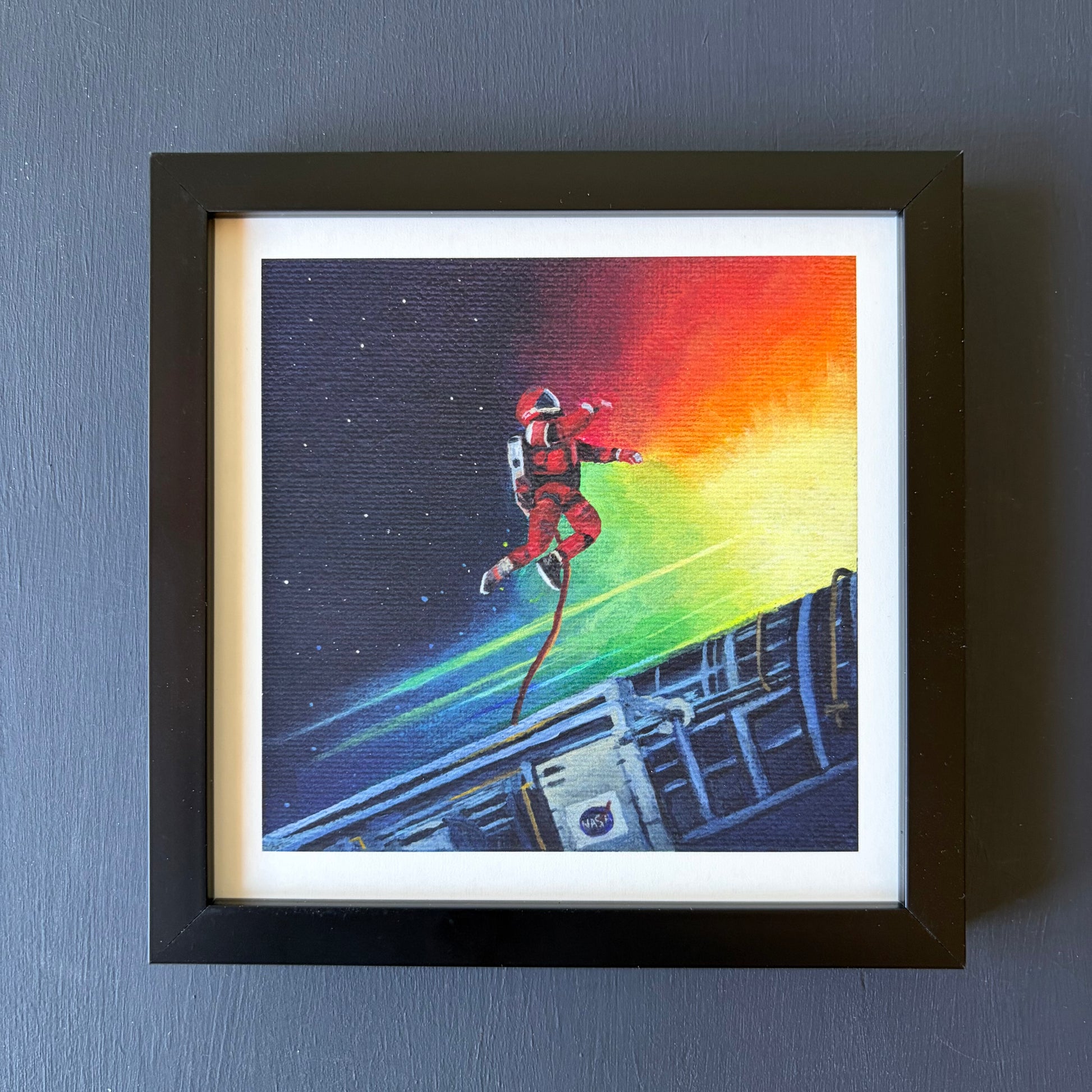 Mini print in black frame of an astronaut tethered to a spaceship with a colorful background