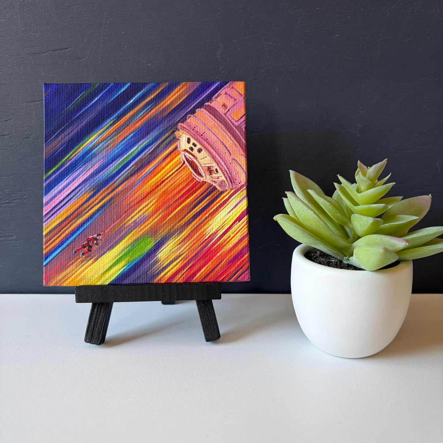 mini painting on easel of a tiny astronaut and spaceship with a background of colorful streaks