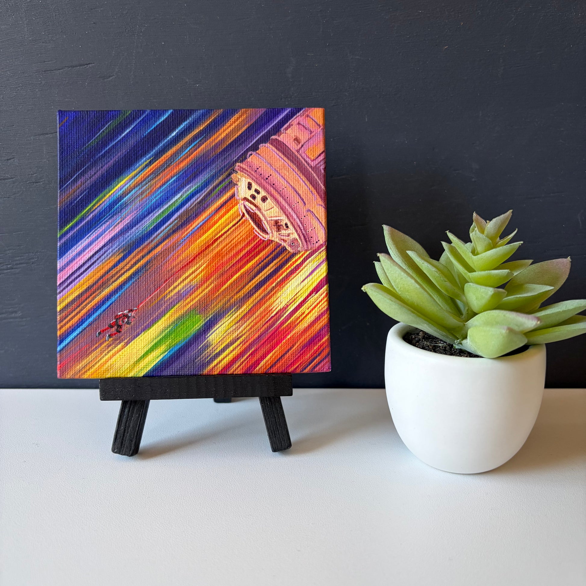 mini painting on easel of a tiny astronaut and spaceship with a background of colorful streaks