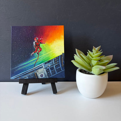  mini painting on easel of an astronaut tethered to a spaceship with a colorful background