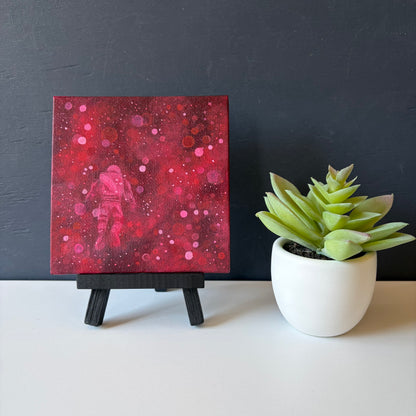 Mini painting on easel of an astronaut floating surrounded by red and pink bokeh 