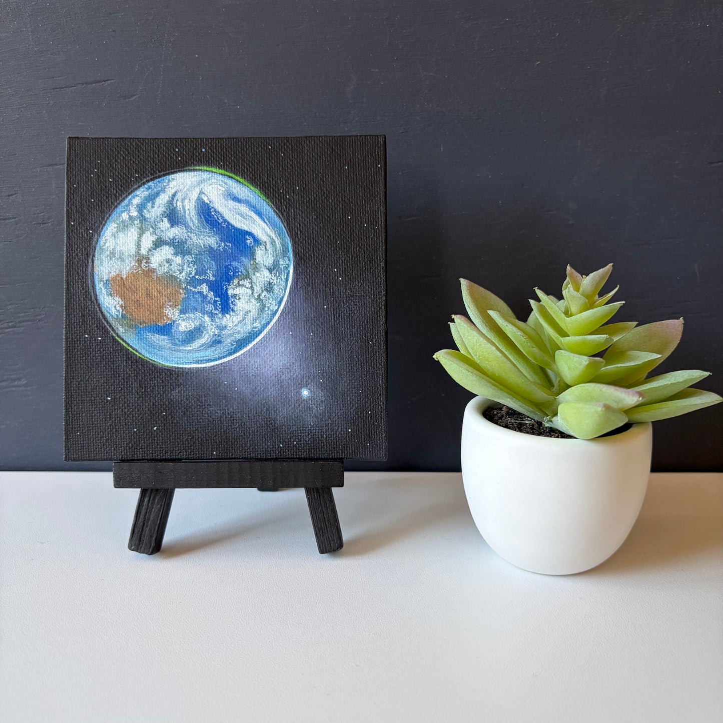 mini painting on easel of Earth from space, Venus in the bottom right, and a black background with a few stars.