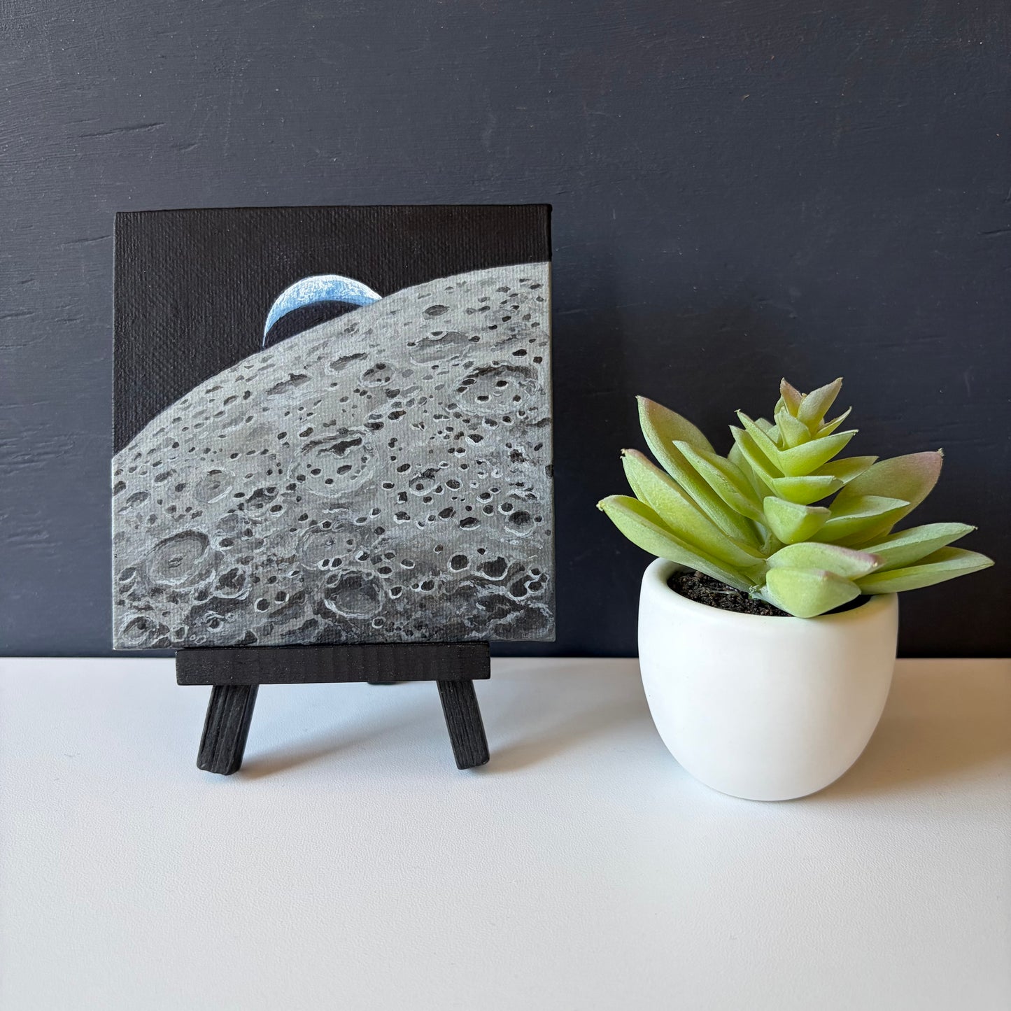 Mini painting on easel of the lunar surface in the foreground and a crescent Earth setting behind the Moon