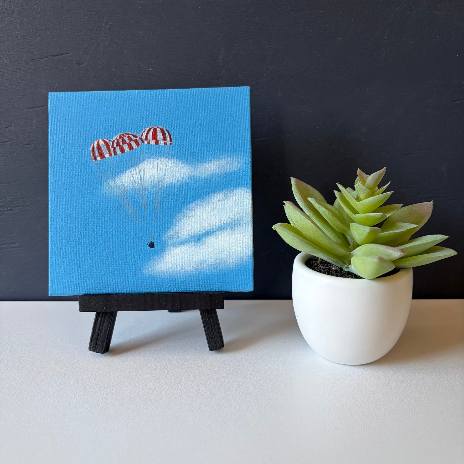 mini painting on easel of 3 red and white striped parachutes descending against a blue sky with clouds