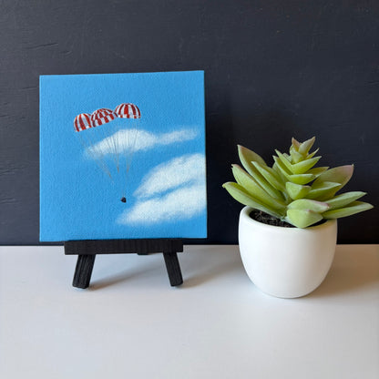 mini painting on easel of 3 red and white striped parachutes descending against a blue sky with clouds