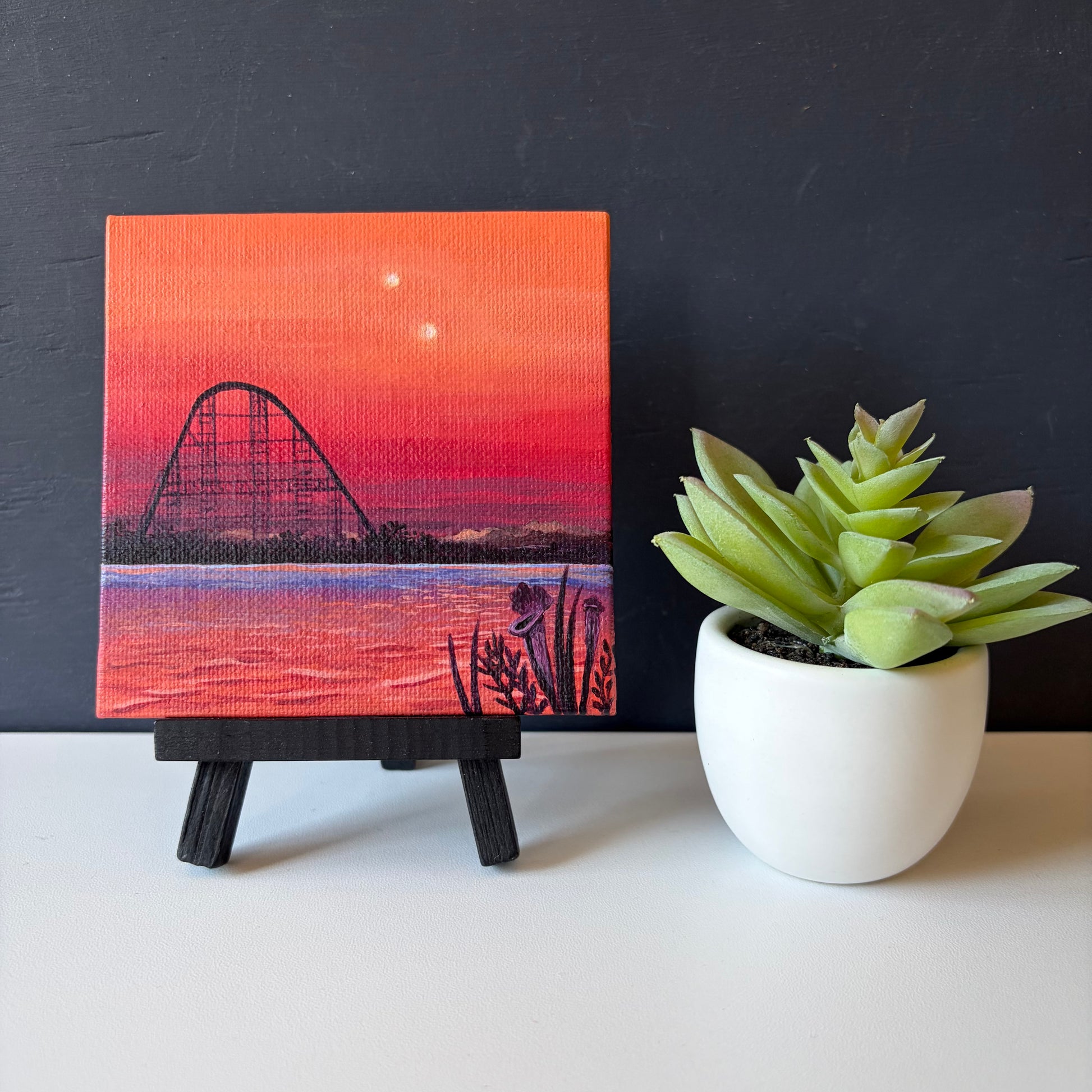 Mini painting on easel. Roller coaster silhouette against a pink and orange sunset sky with water and black plants in the foreground.