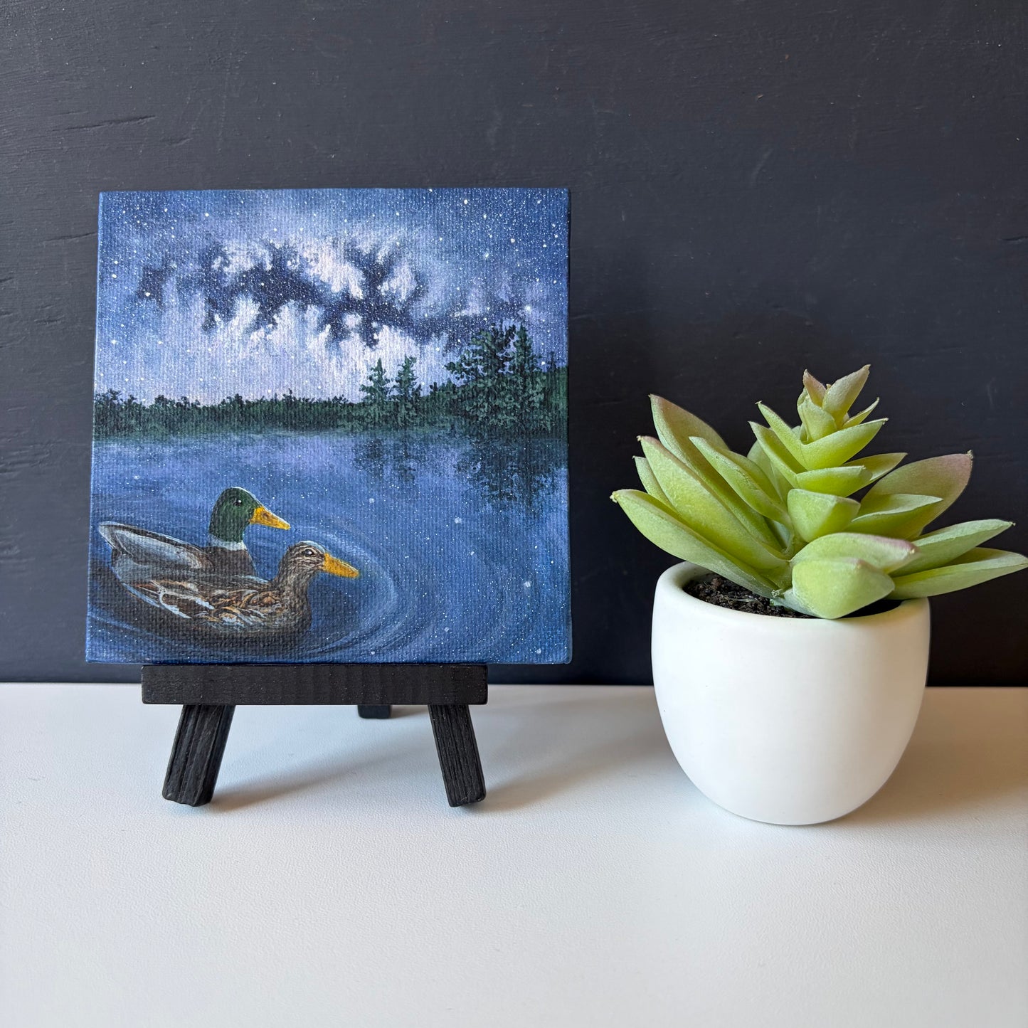 Mini painting on easel. Two ducks swimming on a lake under a starry night sky with trees in the background.