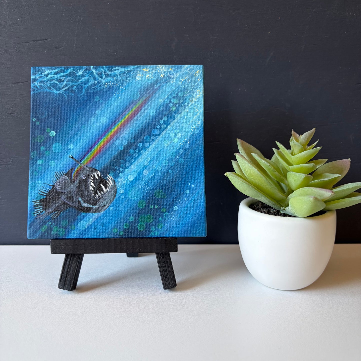 Mini painting on easel of an anglerfish swimming to the surface with a rainbow