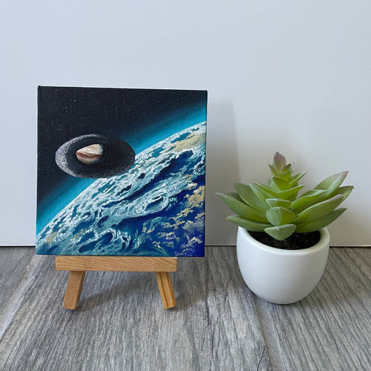 Oval jumpgate orbiting above a blue exoplanet with clouds. The mini painting is on an easel next to a fake plant.