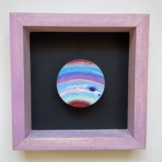 Clay disk painted like Jupiter in pastel colors mounted in a light purple shadowbox frame with black background