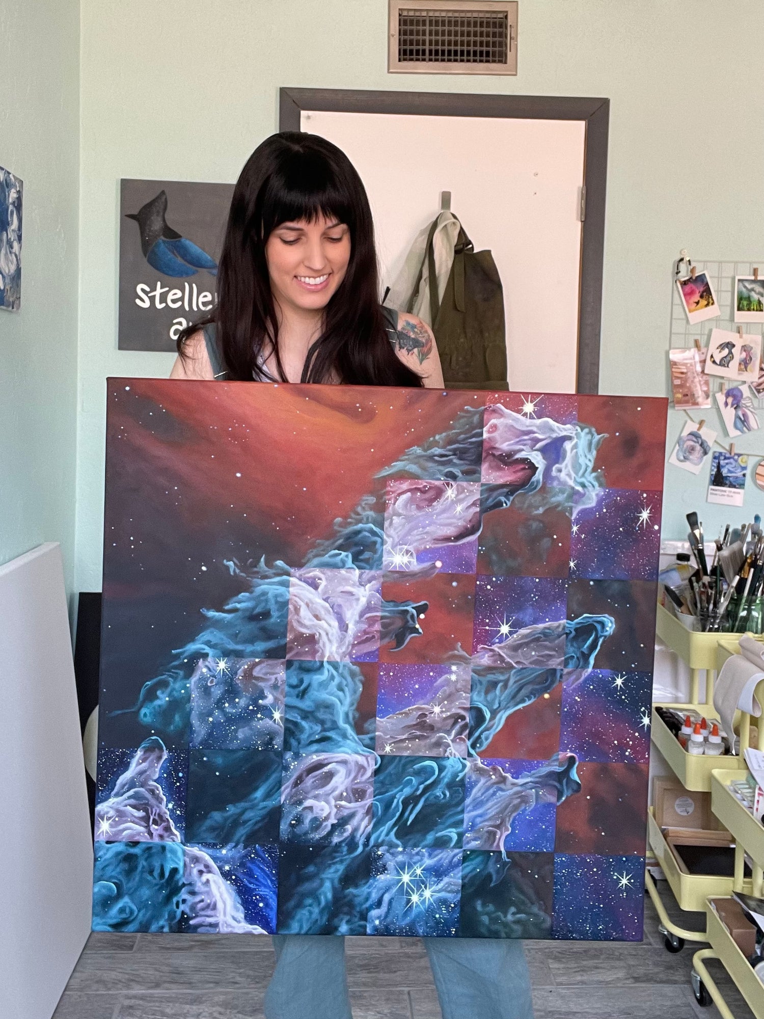 Laci holding a square oil painting of the Pillars of Creation. The painting has a partial checkerboard pattern. The orange squares and blue-grey nebula are how the Pillars look in mid-infrared wavelengths of light. The vibrant purple squares add in shorter, near-infrared wavelengths of light and thousands of stars.