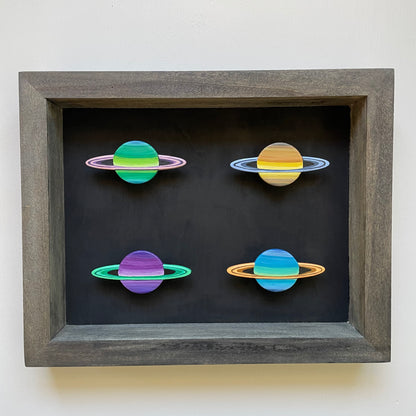 Four mini Saturns painted in vivid colors of green, yellow, purple, and blue mounted in a dark grey shadowbox with black background