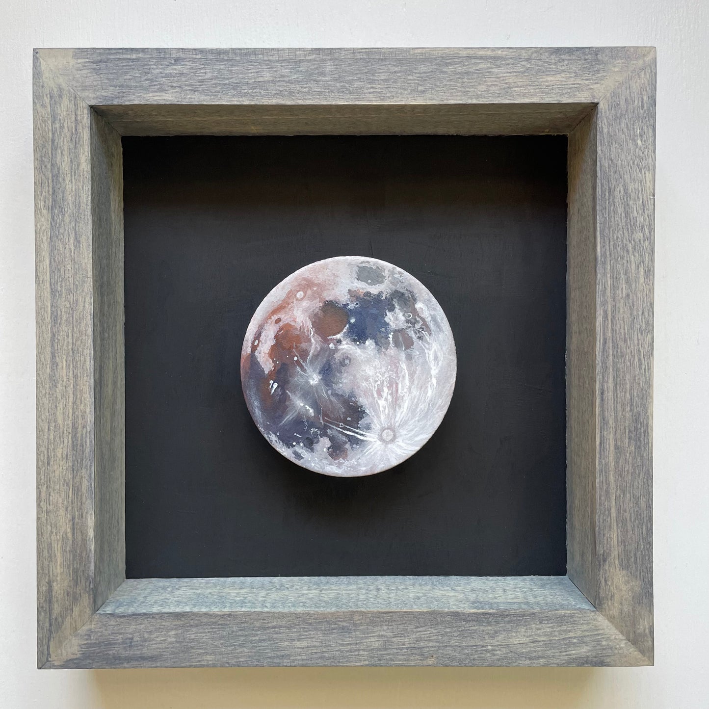 Clay disk painted like Earth's Moon with enhanced colors mounted in a grey stained shadowbox frame with black background