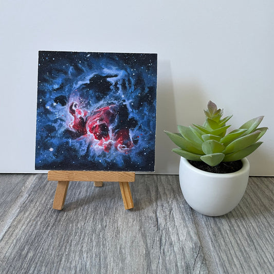 Mini print of a blue nebula with a bright reddish-orange center. The print is sitting on an easel next to a fake plant.