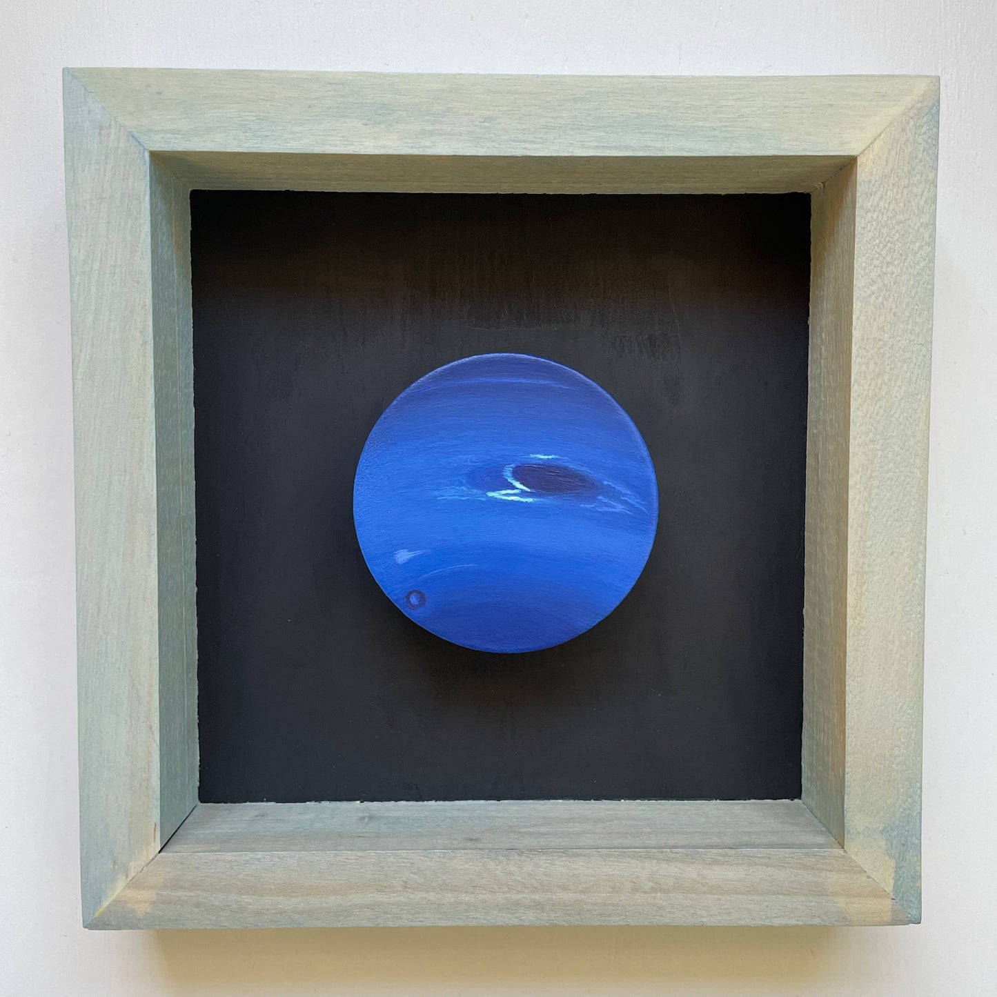 Clay disk painted like Neptune mounted in light blue shadow box with black background