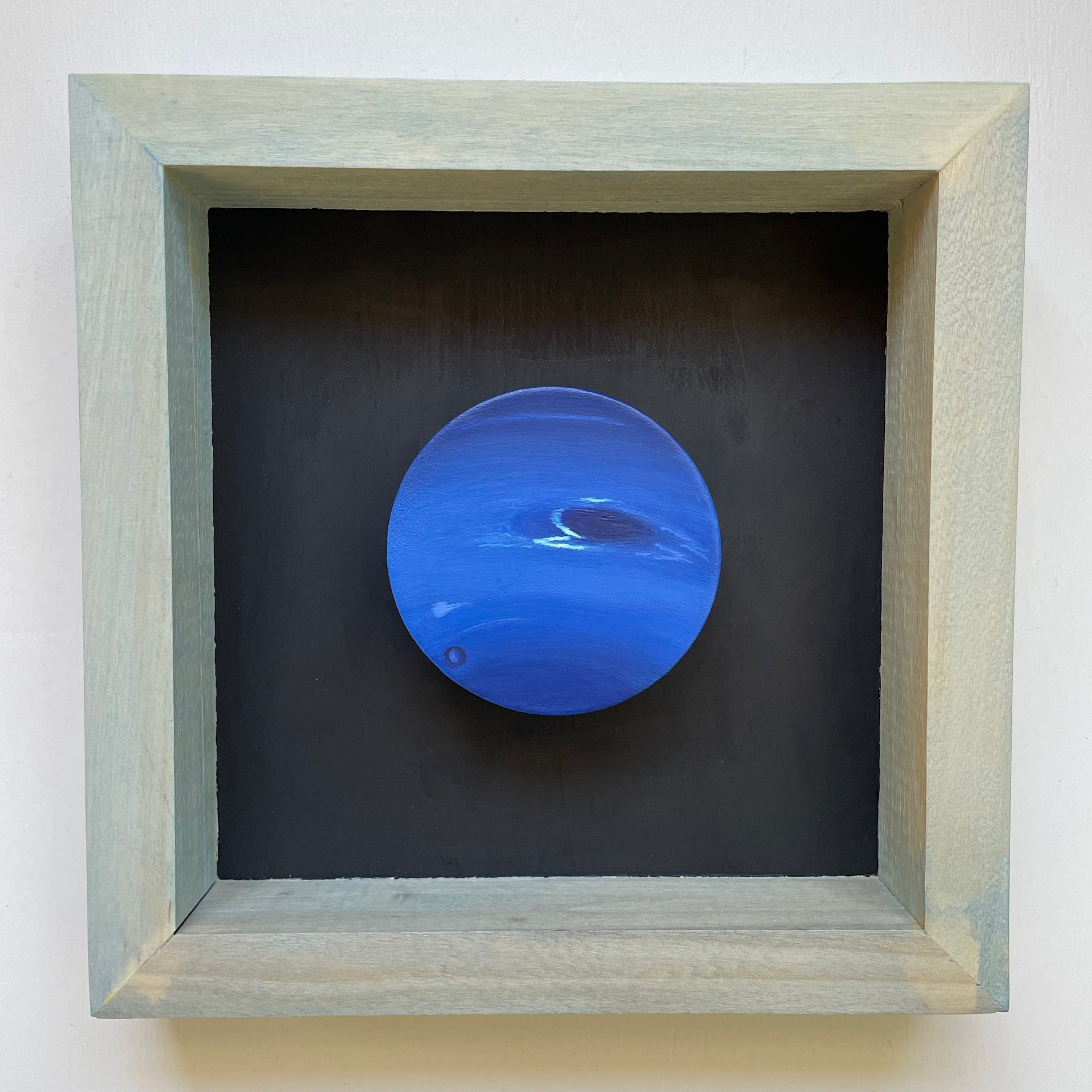 Clay disk painted like Neptune mounted in light blue shadow box with black background