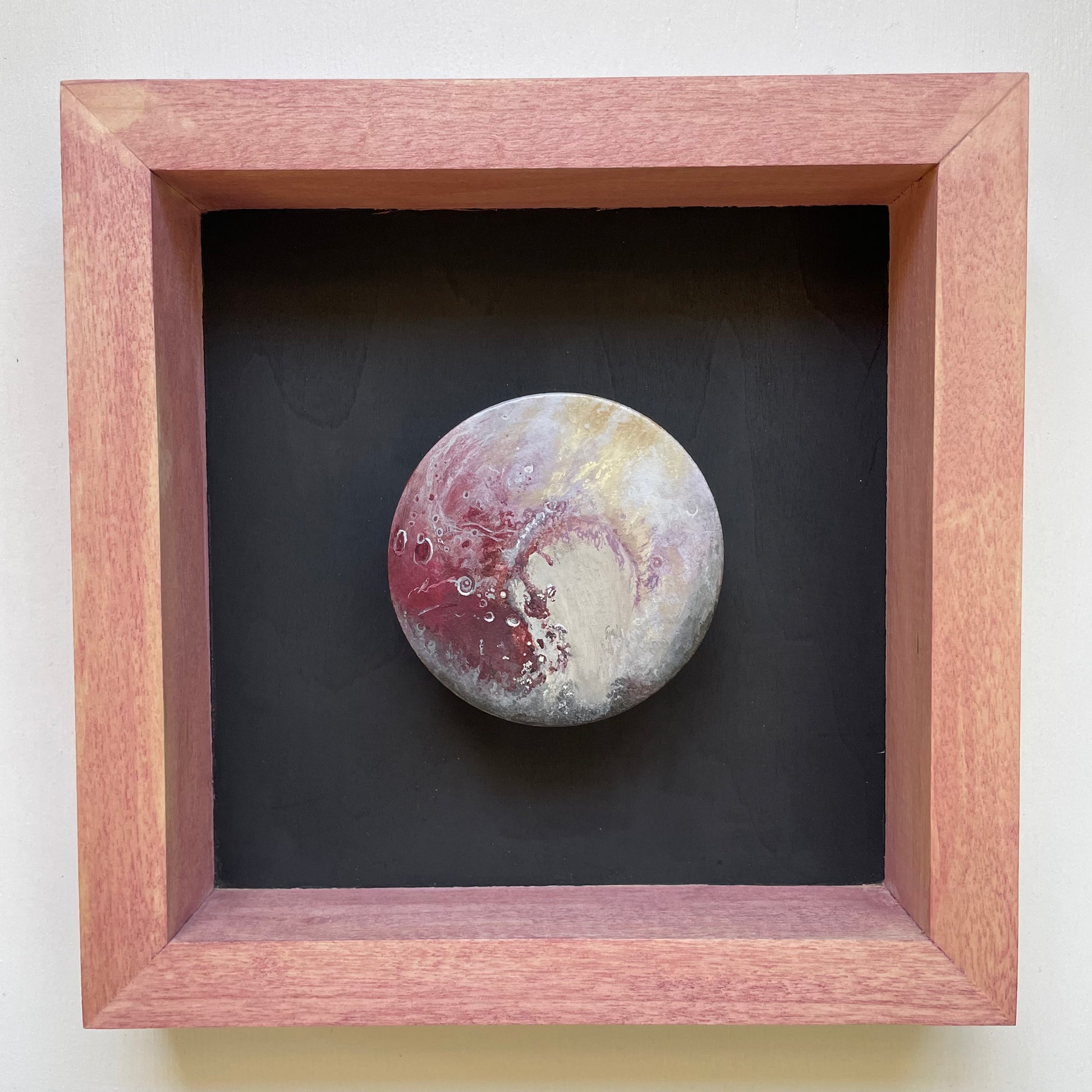 Clay disk painted like Pluto mounted in light red shadowbox frame with black background