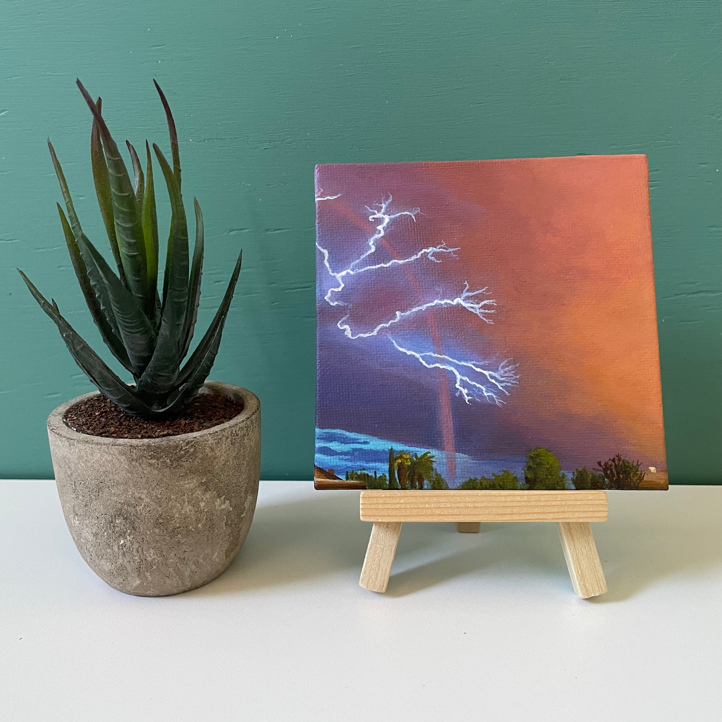 Mini painting of the sky on easel next to fake plant