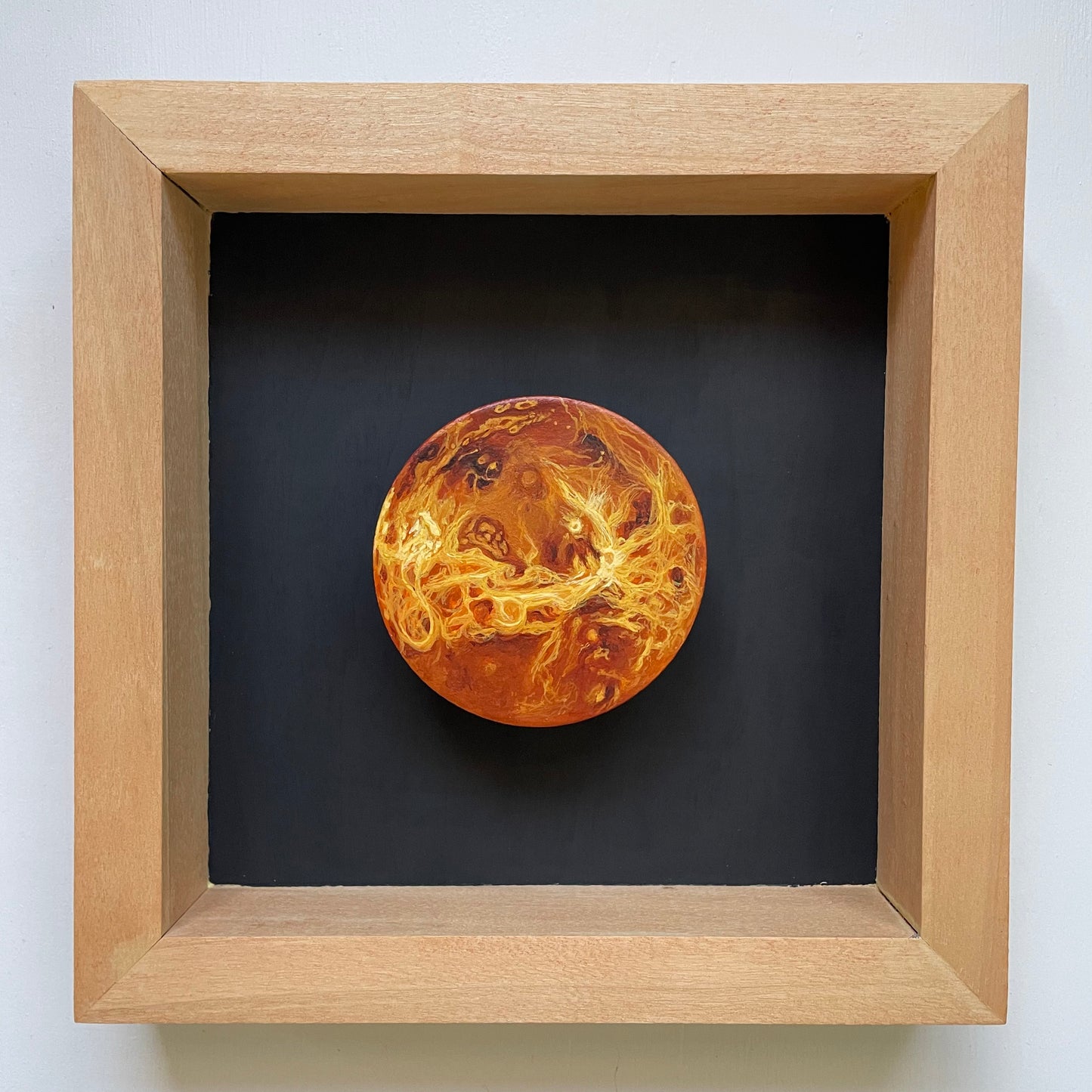 Clay disk painted like Venus mounted in orange stained wood shadowbox with black background